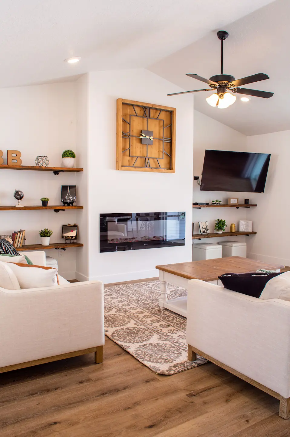 Living room feature wall with a linear electric fireplace, floating wood shelves, and an angled wall-mounted TV; neutral décor with sofa and coffee table.