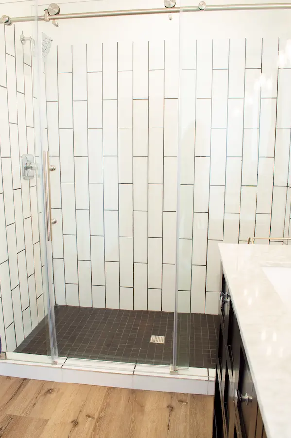 Walk-in shower with vertical white subway tile and glass door; brushed-nickel handle, vanity counter at right.